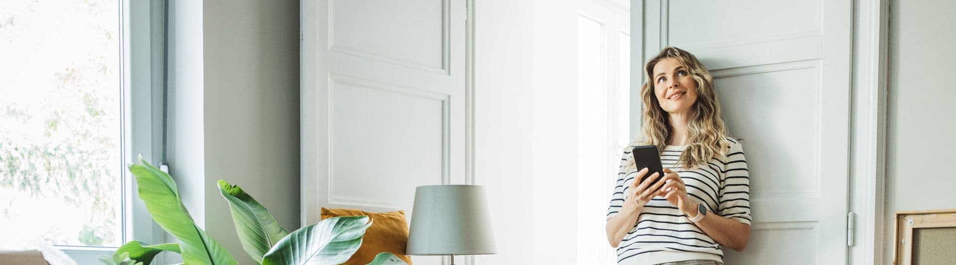 Person står i et lyst rom og holder en mobiltelefon, med bordlampe og grønn plante i bakgrunnen.