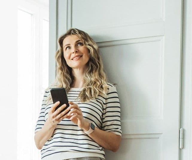 Person står i et lyst rom og holder en mobiltelefon, med bordlampe og grønn plante i bakgrunnen.