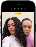iPhone 18 Pro i fargen svart vises forfra, to modeller poserer for kameraet, for å vise Center Stage-frontkameraet